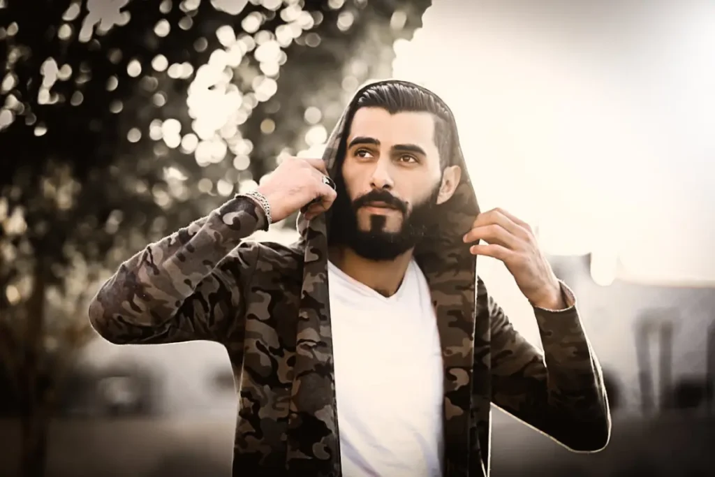 A bearded man with well-groomed hair wearing a hooded jacket in an outdoor